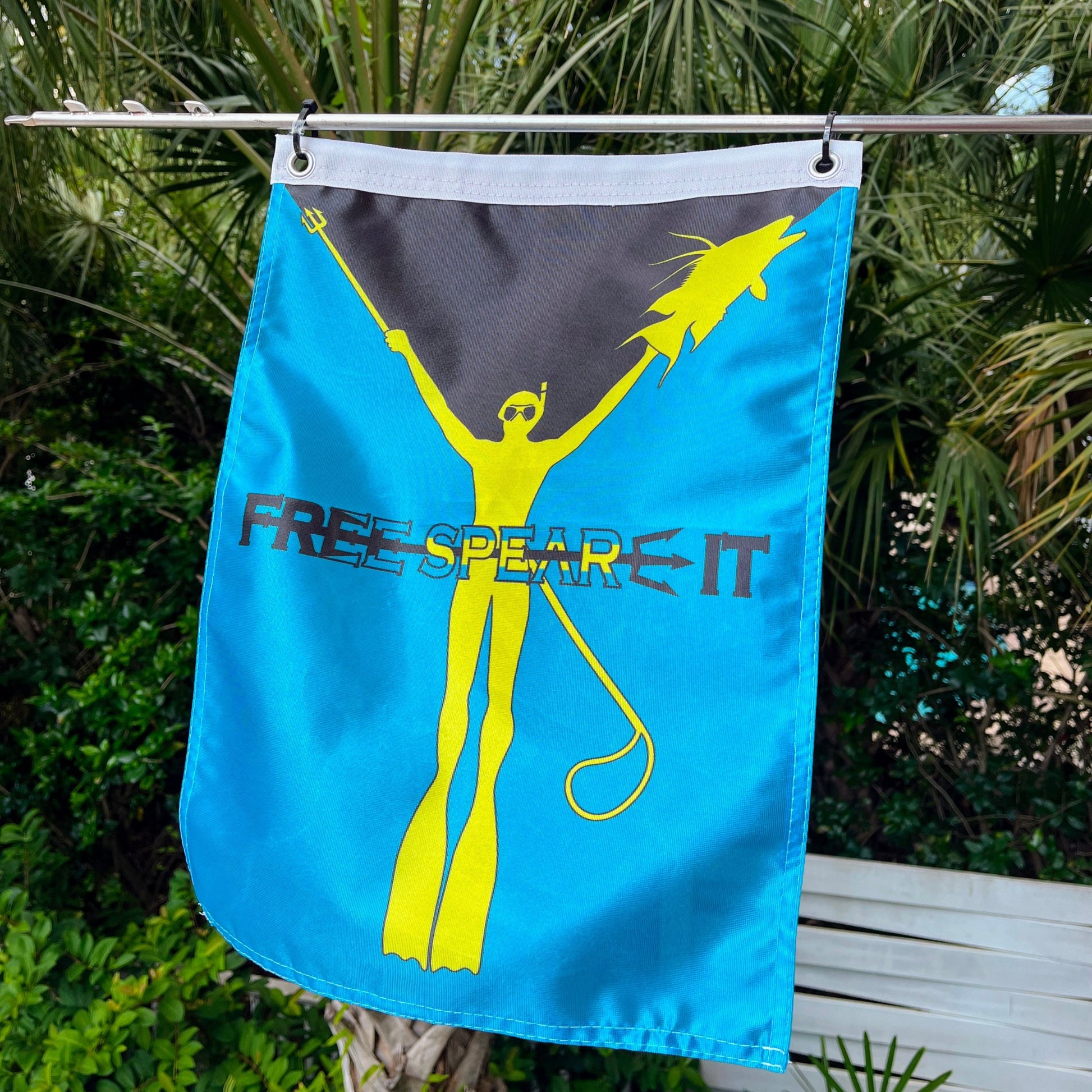 Free Spear-It Bahamas Bounty Flag hanging outdoors on a metal rod, featuring a bright yellow freediver holding a speargun and fish against a vibrant blue background, with palm trees and greenery in the backdrop.