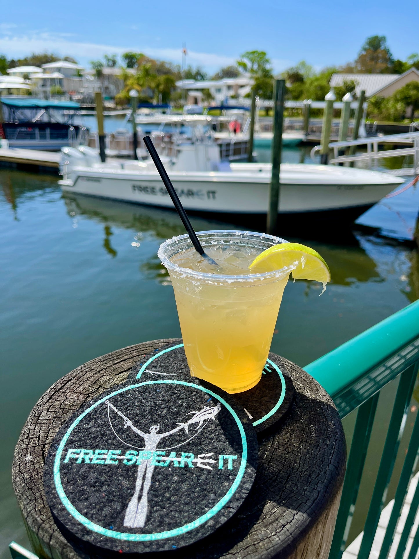 Lifestyle image featuring Free Spear-It Eco Coasters crafted from recycled wetsuits by Lava Rubber. Set against a scenic marina with a Free Spear-It boat in the background, the coasters hold a refreshing beverage, showcasing their durability and eco-friendly design. Perfect for outdoor adventures, boating, and sustainable living enthusiasts.