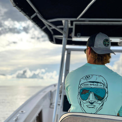 Lifestyle shot of a man at the helm of an open fisherman boat wearing the Free Spear-It Deep Reflections Long Sleeve UPF 50+ Solar Performance Shirt in seagrass green, featuring a bold graphic design of a freedive spearfisherman’s face with a mirrored dive mask reflecting a speargun and hogfish. This lightweight, sun-protective shirt is perfect for spearfishing enthusiasts who value comfort and style during their adventures. Available now at FreeSpear-It.com.