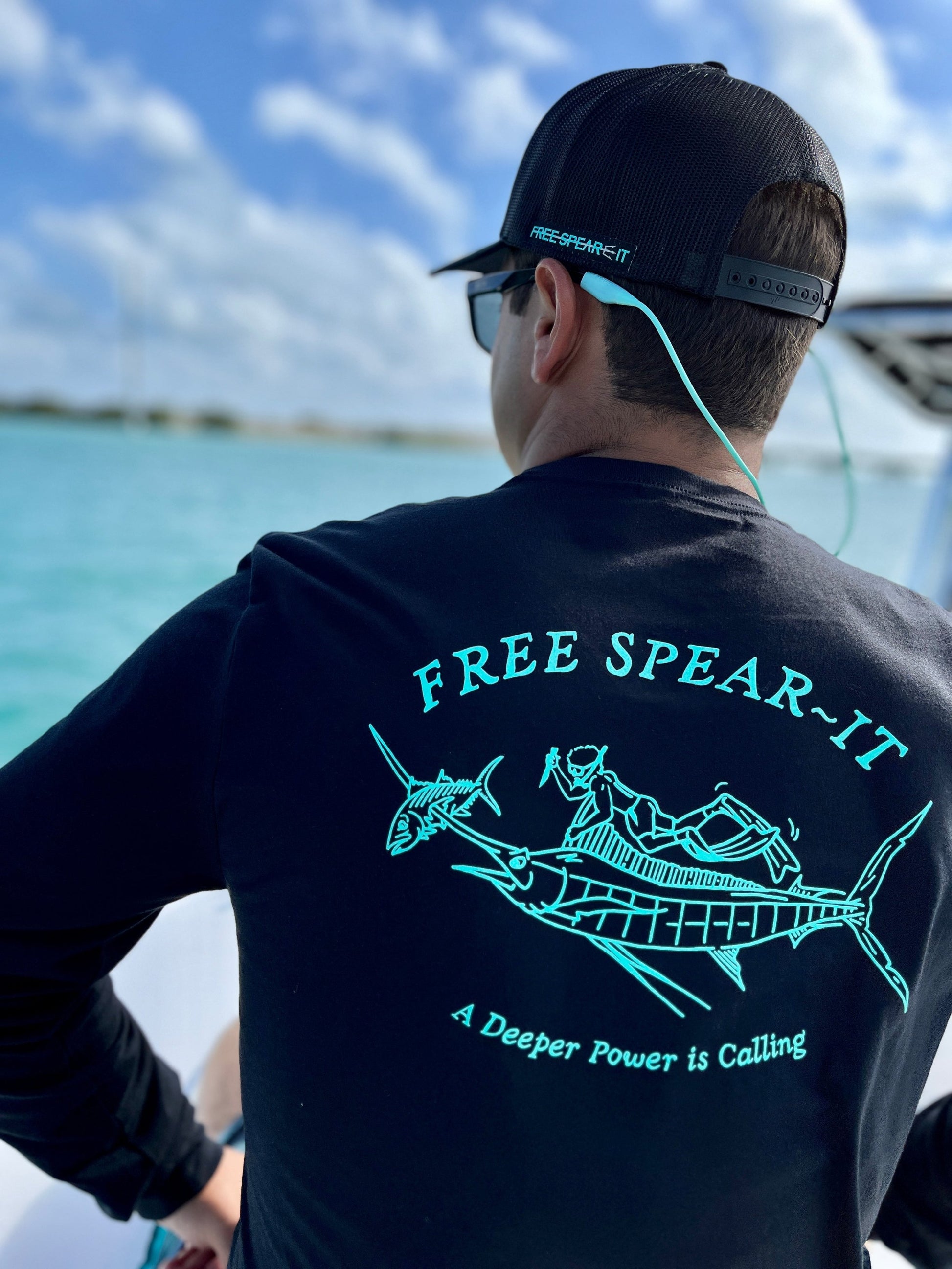 Back view of a person sitting on a boat on the water wearing the Free Spear-It Marlin Mayhem Long Sleeve T-Shirt in black, featuring a detailed graphic in aqua of a freediver swimming with a marlin that has speared a fish on its bill.  The design includes the words Free Spear-It and the slogan "A Deeper Power is Calling." 