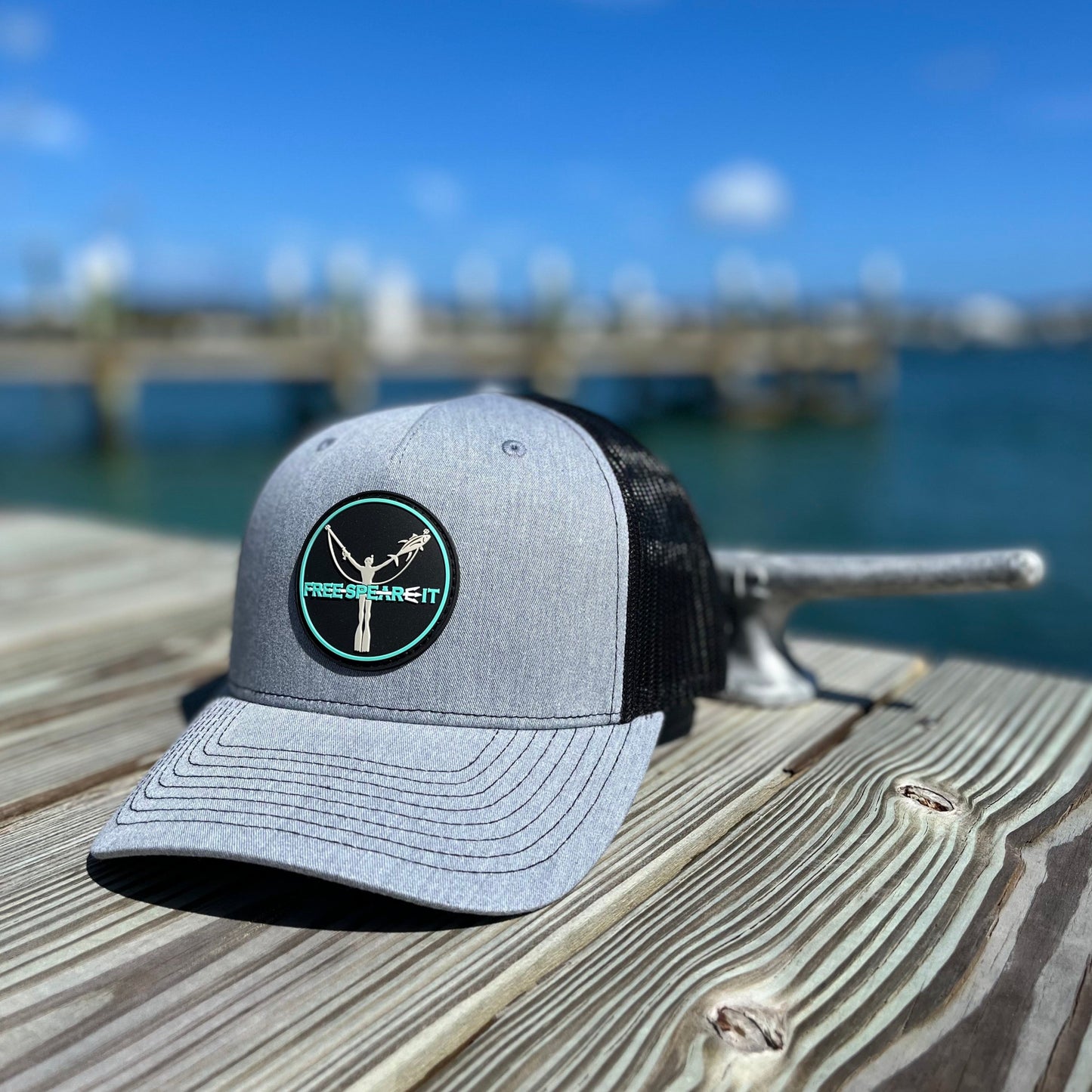 Lifestyle photo of the Free Spear-It PVC Logo Snapback Trucker Hat in heather grey and black, featuring a durable PVC patch with a freediver design. Captured dockside with a marina backdrop, this Richardson five-panel hat is perfect for spearfishing, boating, and outdoor adventures.
