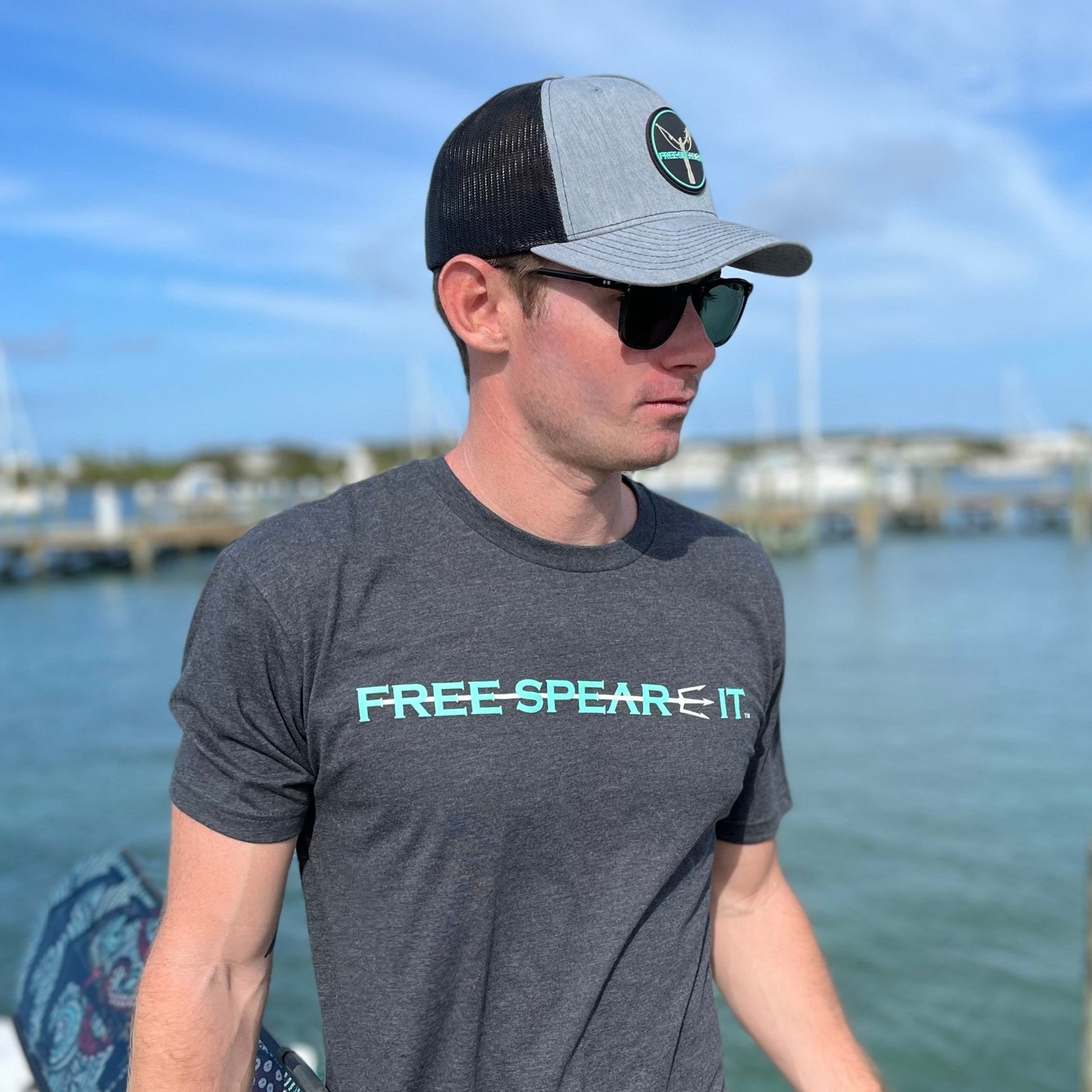 Man wearing a charcoal Free Spear-It t-shirt and PVC logo snapback hat, holding freedive fins and a pole spear while standing on a dock. This photo captures the spearfishing lifestyle and showcases comfortable, ocean-ready apparel for freedivers and outdoor adventurers.