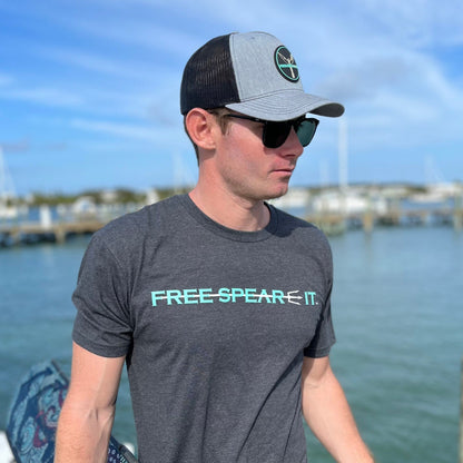 Man wearing a charcoal Free Spear-It t-shirt and PVC logo snapback hat, holding freedive fins and a pole spear while standing on a dock. This photo captures the spearfishing lifestyle and showcases comfortable, ocean-ready apparel for freedivers and outdoor adventurers.