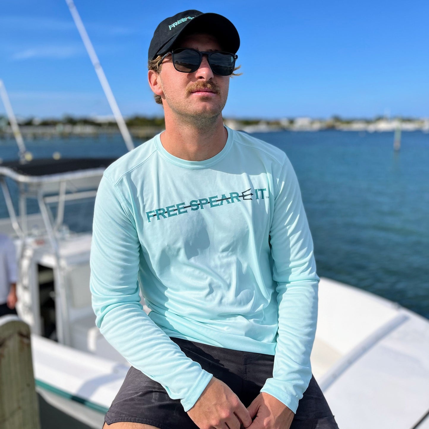 A man wearing the Free Spear-It Deep Reflections Long Sleeve UPF 50+ Solar Performance Shirt in seagrass and a Free Spear-It hat, seated on a dock with a boat and ocean in the background. This sun-protective, lightweight spearfishing shirt combines comfort and performance, making it an ideal choice for spearfishing, boating, and outdoor adventures. Shop premium spearfishing gear and apparel at FreeSpear-It.com.